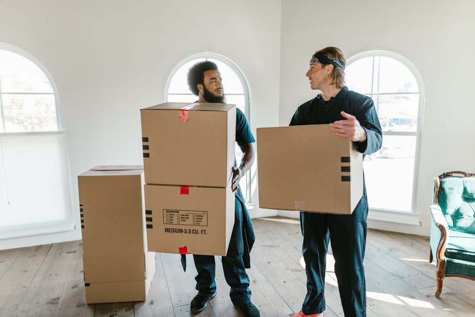 A man wearing a dark blue work jumpsuit and a black bandana with white patterns on his head is inside a property, standing next to a cardboard box filled with various items. He is holding a clipboard