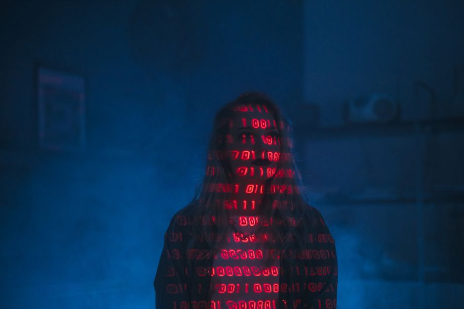 A person with long hair standing indoors in front of a dark blue wall, with their face and upper body illuminated by red digital binary code projections that display sequences of 0s and 1s on their cl