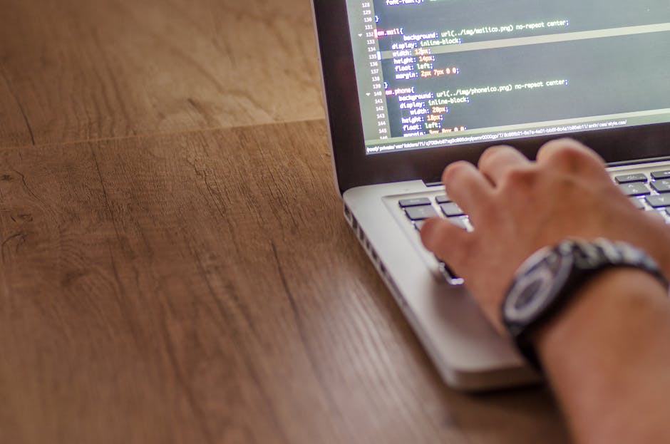 The image shows a person using a laptop placed on a dark table, with their hands operating the keyboard. The laptop screen displays a dark-themed code editor with lines of colorful programming code, i