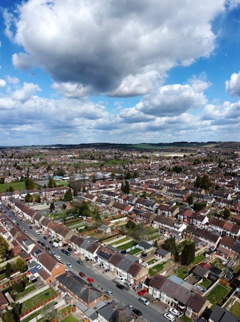 Aerial view of a residential street showing a moving truck parked on the driveway of a modern detached house, which has a front garden with a neatly trimmed lawn and a paved driveway. The street featu