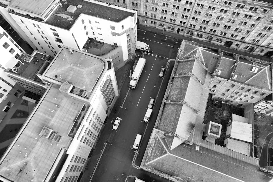 A black and white aerial photograph showing a narrow city street surrounded by tall residential and commercial buildings with flat roofs. Several white vans and smaller vehicles are parked along both 
